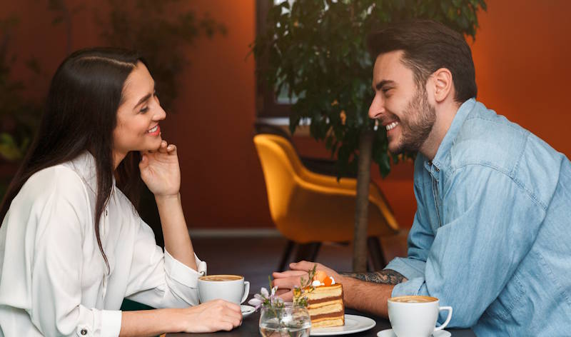  Couple souriant et complice partageant un café et un gâteau dans une ambiance chaleureuse, illustrant l’écoute, la tendresse et la communication dans une relation amoureuse.